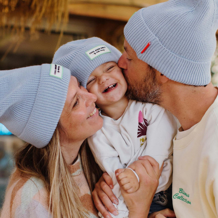Family wearing matching light blue beanies with visible hello hossy brand logos, smiling outdoors.