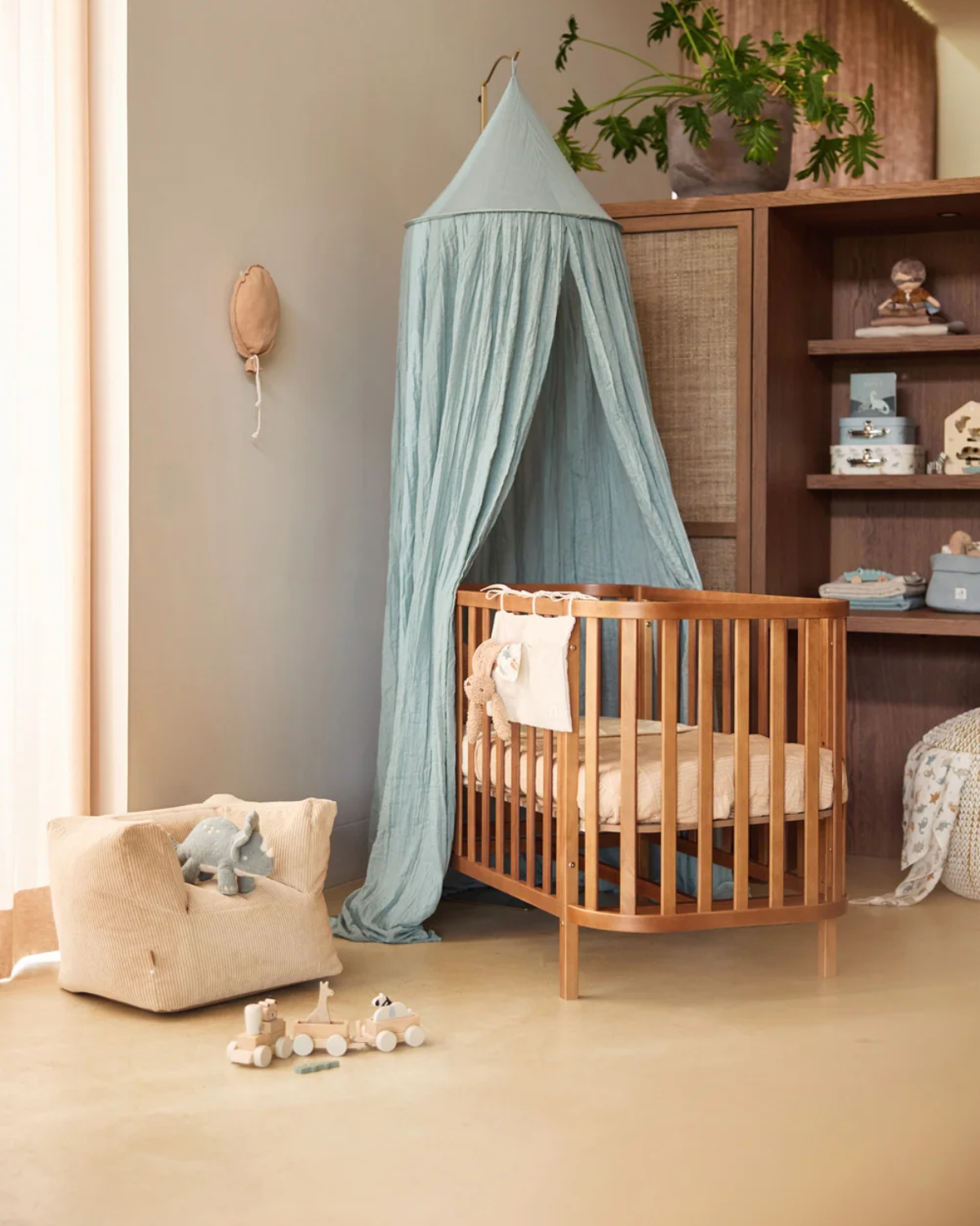 A wooden baby crib with a light blue canopy tent in a softly styled nursery. The crib is made of warm medium-brown wood and is dressed in cream bedding.